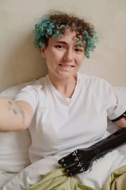 Protez kollu, gülümseyen kıvırcık kadın yatakta selfie çekiyor.