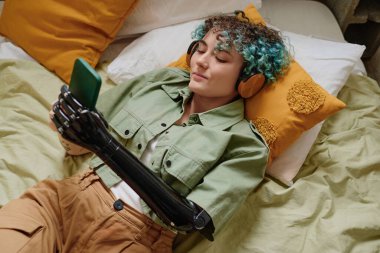 Protez kollu gülümseyen genç bir kadın kulaklıkla müzik dinliyor ve akıllı telefondan sosyal medyayı kontrol ediyor.