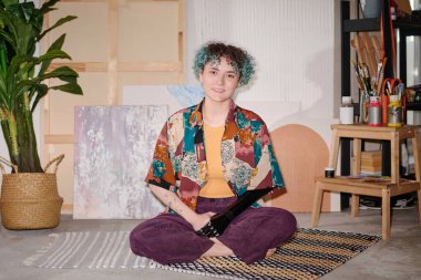 Yaratıcı stüdyoda yerde oturan protez kollu gülümseyen sanatçının portresi.