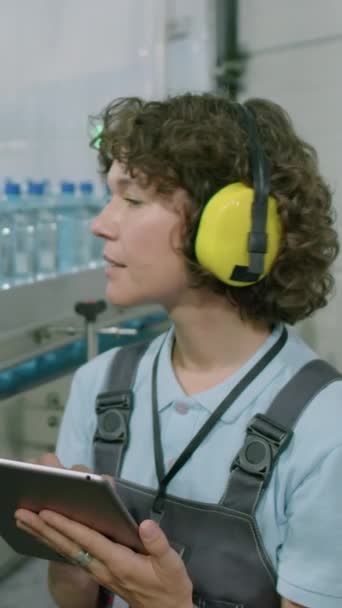飲料工場でデジタルタブレットを使用してコンベヤーにボトルを数える耳プロテクターの女性労働者の垂直ショット — ストック動画