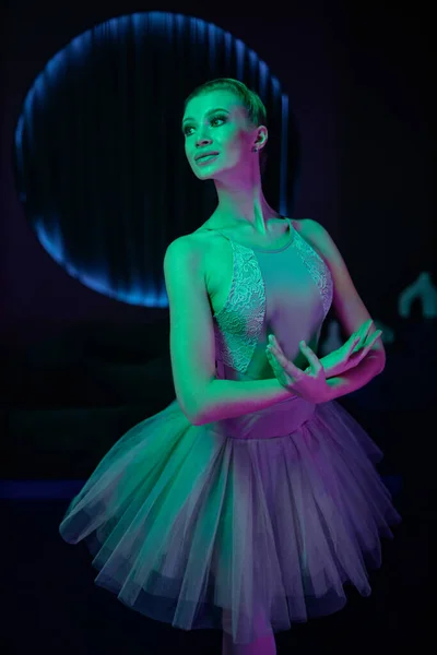 Medium full shot of ballerina posing under neon lights in dark studio