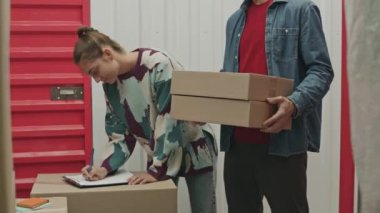 Kadın çalışanın depolama biriminin içerisinden panoya not alıp erkek müşteriye kutuları nereye koyması gerektiğini söyler