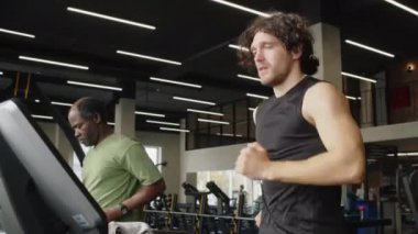 Beyaz tenli genç sporcu ve orta yaşlı bir adamın modern spor salonundaki koşu bantları eğitiminin düşük açılı çekimi.
