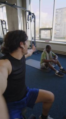 Sporcunun birini videoda ararken ve spor salonunda spor yaparken arkadaşının yerde kıvrımlar yaptığını söylerken dikey görüntüsü.