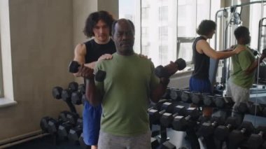 Orta yaşlı erkek çalıştırıcının spor salonunda dambıllarla egzersiz yaparken orta yaşlı erkek duruşunu düzeltmesi.