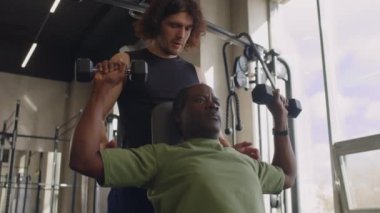 Düşük açılı orta yaşlı amatör sporcunun dambılları kaldırırken erkek koçun arkada durup spor salonunda yardım ederken.