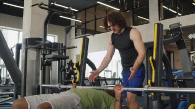 Orta boylu, orta yaşlı, spor salonunda halter kullanarak göğüs egzersizi yapan Afrikalı Amerikalı bir adama yardım eden Kafkas antrenörü.