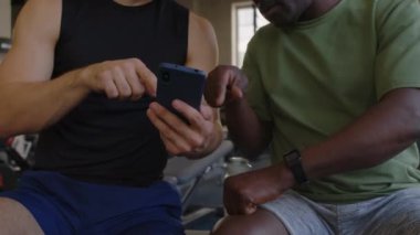 Spor salonunda antrenman yaptıktan sonra spor salonunda dinlenirken erkek koçun cep telefonuna fitness uygulaması önermesi.