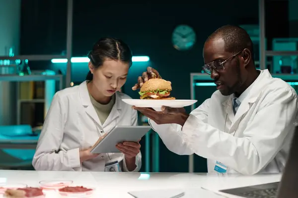 Black scientist examining hamburger with lab-grown meat, his colleague working on tablet nearby