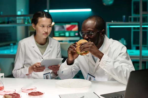 Black scientist smelling hamburger with artificial meat, his asian colleague sitting nearby