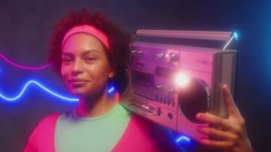 Yüzünde retro teyp olan gülümseyen zenci kadının orta boy portresi. Stüdyoda neon ışıklarla kameraya bakıyor.