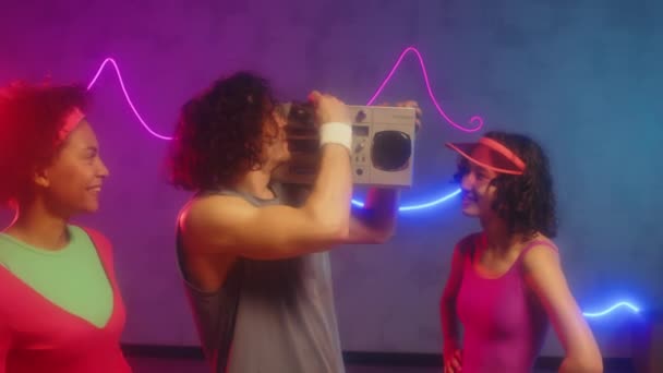 ダンスやエアロビクスのクラスでレトロテープレコーダーにカセットをロードする男性トレーナーのミディアムショット 女性の友人がネオンスタジオで音楽を踊る — ストック動画