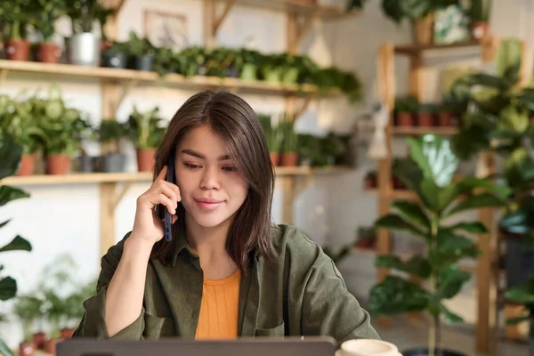 Young plant store manager talking on mobile phone while browsing laptop sitting at desk at store