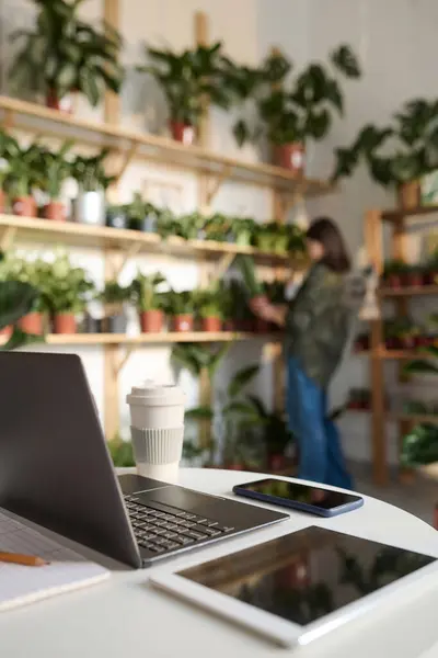 Electronic devices displayed on desk in plant store, worker arranging plants on background