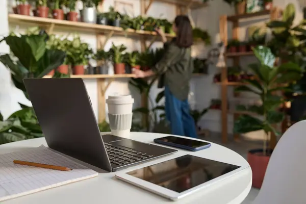 Electronic gadgets on desk in plant store, worker checking plants on shelves on background