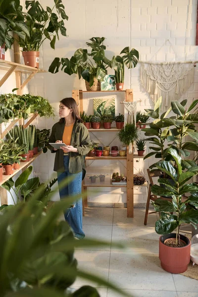 Young plant shop employee checking potted plants on shelves with tablet in her hands