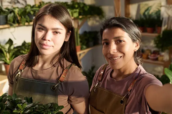 Selfie of two female plant shop workers standing at their workplace and smiling at camera together