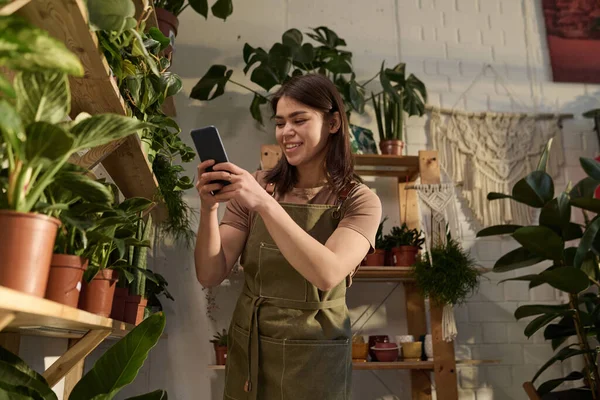 Medium full shot of young cheerful plant shop worker filming racks with plants on her smartphone