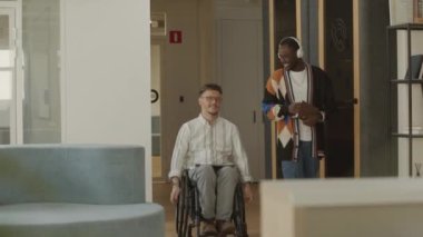 Genç Kafkasyalı işadamı, tekerlekli sandalyeye biniyor ve iş günlerinde modern ofisin içinde yürüyen Afrikalı Amerikalı erkek iş arkadaşıyla sohbet ediyor.