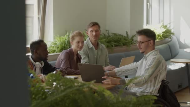 ノートパソコンでビジネスプロジェクトを見せて 非公式のオフィスミーティング中に多民族の同僚と話し合う車椅子の若い男 — ストック動画