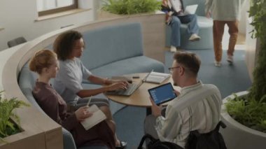 Tekerlekli sandalyedeki bir adamın dijital tablet kullanırken ve iki farklı etnik gruptan kadın meslektaşıyla yeşil bitkilerle süslenmiş modern çalışma odasında konuşurken yüksek açılı görüntüsü.