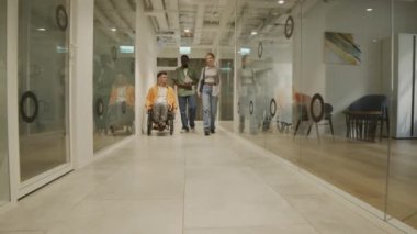 İki ofis çalışanı ve tekerlekli sandalyedeki işadamı koridorda birlikte yürüyorlar ve sohbet ediyorlar.