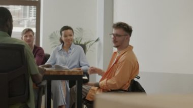 Tekerlekli sandalyedeki genç proje yöneticisi, gayri resmi ofis toplantısında çok ırklı iş ekibiyle fikirleri tartışıyor.