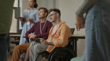 Tekerlekli sandalyedeki adam dijital tablet tutuyor ve kurumsal eğitim etkinliği sırasında konuşmacı sunumu sırasında sorular soruyor.