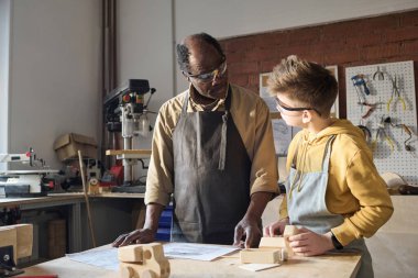 Siyah kıdemli marangozun portresi küçük çocuğa atölyede ahşap işçiliği öğretiyor ve planlara bakıyor.