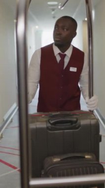 Üniformalı ve beyaz eldivenli genç Afrikalı komi otel koridorunda yürürken ve bagaj arabasında bavul taşırken dikey bir fotoğraf.