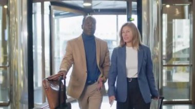 Afro-Amerikalı işadamı ve Kafkasyalı iş kadını giriş koridorunda bavullarıyla yürüyorlar ve otele birlikte varırken sohbet ediyorlar.