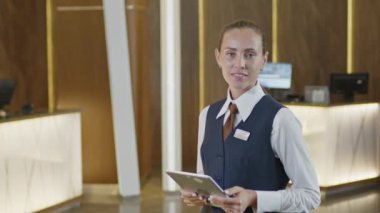 Resmi kıyafetli, elinde dijital tabletle genç bir bayanın orta boy fotoğrafı ve otelde kameraya gülümseyerek poz vermesi.