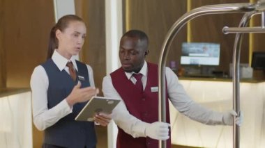 Dijital tablet kullanan ve genç Afrikalı Amerikalı kapıcıya iş görevini açıklayan bir kadın otel müdürü onu dinliyor ve bagaj arabasıyla uzaklaşıyor.