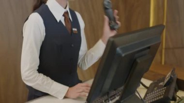 Formalite icabı çalışan genç bayan resepsiyon müdürü telefonu kaldırıyor, müşteriyle konuşuyor ve iş saatlerinde bilgisayar kullanıyor.
