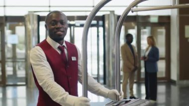 Beyaz eldivenli, üniformalı, oteldeki bagaj arabasıyla kameraya gülümseyerek poz veren genç Afrikalı Amerikalı hamal portresi.