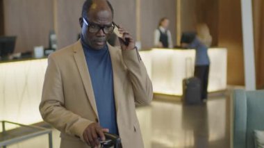 Siyah işadamının bavulunu otel lobisinde dikerken ve cep telefonundan arama yaparken çekimini yap.