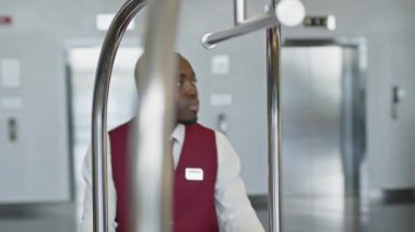 Genç Afrikalı Amerikalı hamalın otelin lobisindeki çıkış kapısına doğru giden valiz arabasını çekerken.
