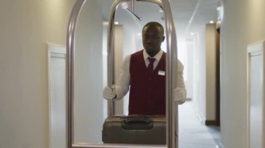 Genç Afro-Amerikan üniformalı hamal otel koridorundan geçiyor ve bagaj arabasını itiyor.