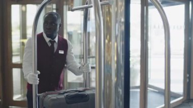 Genç Siyah komi üniformalı ve eldivenli valiz arabasını otel lobisine itiyor.