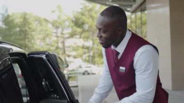 Siyah üniformalı genç hamal arabanın kapısını açıyor ve karşılayan kadın otele taksiyle geliyor.