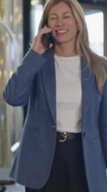 Dikey olarak, gülümseyen kadının bavullarıyla otelin lobisinde yürürken ve iş gezisi sırasında cep telefonuyla konuşurken görüntüsü.