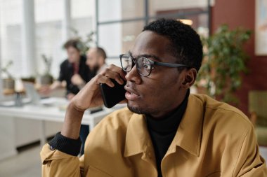 Siyah ofis çalışanı iş yerinde akıllı telefondan konuşuyor, meslektaşları arka plan üzerinde çalışıyor.