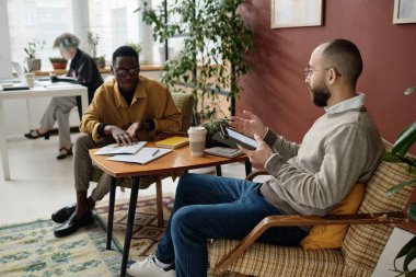 Çift ırklı erkek iş arkadaşları ofiste masada oturan bilgili belgeleri tartışıyor.