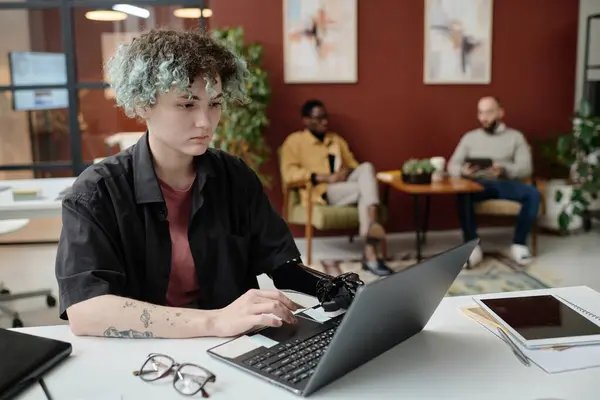 Young girl with prosthetic arm browsing laptop at office, her male colleagues sitting on background