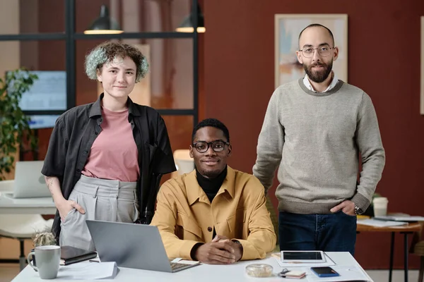 Two biracial male coworkers and their female colleague posing by office desk smiling at camera