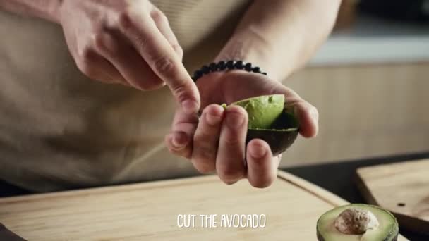 シェフの熟したアボカドの手を切断するショーを調理するためのビデオチュートリアルでは キッチンテーブルで果物をスクープしてスライスする方法を示し 下に表示されるステップ説明の字幕 — ストック動画