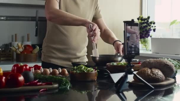 男子在平底锅里搅拌食物和在厨房桌上用手机拍摄烹调过程的中段镜头 — 图库视频影像