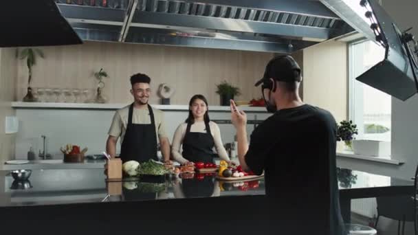 食材とレシピを伝えるキッチンテーブルに立って若い男性と女性シェフが主催するテレビ番組を撮影するカメラマン — ストック動画