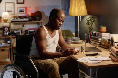 Engelli kıdemli siyahi adam evde oturmuş ilaç organizatörünü dikkatle inceliyor.