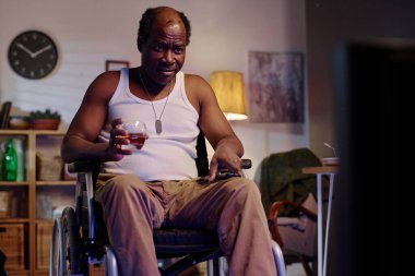 Mutlu, emekli Afro-Amerikalı, tekerlekli sandalyede televizyon seyrediyor ve alkollü içki içiyor.